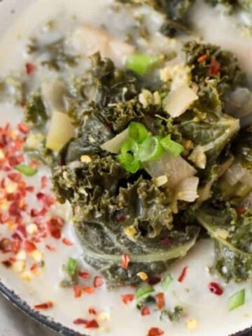 A bowl of creamy soup topped with kale, chopped green onions, and red pepper flakes. A spoon rests on the bowl's edge. The dish sits on a textured surface.