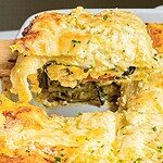 A close-up of a slice of cheesy spinach and artichoke casserole being lifted from the dish with a spatula. Reminiscent of butternut squash lasagna, this casserole is topped with melted cheese and garnished with parsley.