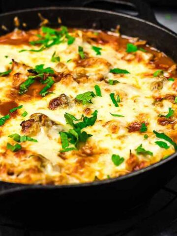 A cast iron skillet filled with baked pasta topped with melted cheese and garnished with chopped parsley. It sits on a wire rack. A small bowl of shredded cheese and a spoon of additional parsley are nearby on the gray surface.