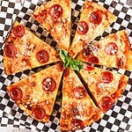 A pepperoni pizza, baked to perfection in a cast iron skillet, is cut into eight slices on a checkered paper. The deep dish creation features a crispy crust with melted cheese, generous pepperoni slices, and sprinkled parmesan. A garnish of fresh basil crowns the center.