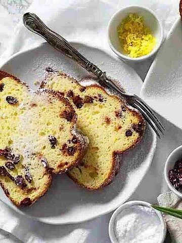 A sliced loaf of sweet bread topped with powdered sugar is on a long white platter. Nearby, two slices rest on a small round plate with a fork. Lemon zest, dried cranberries, and a red-yellow tulip are also visible on the table.