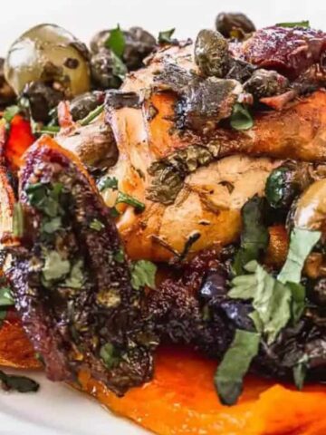 A plate of roasted chicken topped with herbs, olives, and capers lies on a bed of sweet potato wedges reminiscent of classic casseroles. A fork is artfully placed to the right, evoking memories of cooking for years.