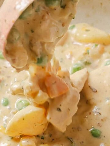 A bowl of creamy stew containing chunks of chicken, peas, corn, and diced potatoes. A ladle lifts a portion of the stew, showing the thick consistency and mixture of ingredients.