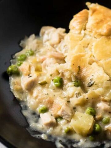 A black bowl contains chicken pot pie with a flaky golden crust. The filling includes chicken chunks, peas, and potatoes in a creamy sauce. A white cloth is partially visible underneath the bowl.