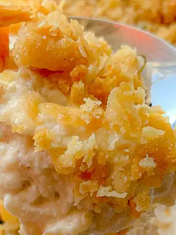 A close-up of a spoon scooping a creamy casserole topped with crushed golden crackers and small herb leaves. The dish is textured with a mix of creamy and crispy elements, suggesting a comforting and rich flavor.