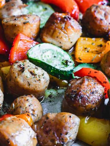 Roasted vegetables and sausages on a baking sheet, featuring red and yellow bell peppers, zucchini slices, and browned sausage pieces. This kitchen favorite is seasoned with herbs, glistening with oil, and perfect for those seeking low-effort recipes.