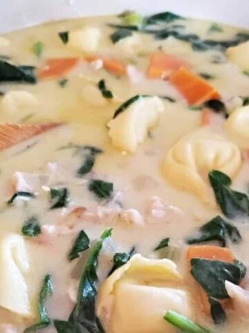 A pot of creamy soup is being stirred with a wooden spoon. The soup contains tortellini, spinach, diced carrots, and pieces of chicken, all in a light-colored broth.