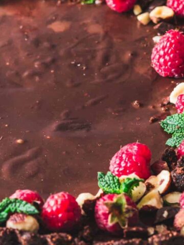 Close-up of a chocolate tart with a smooth surface, topped with fresh raspberries, chopped nuts, chocolate pieces, and mint leaves. The tart is placed on a light-textured surface.
