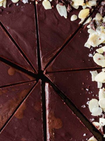 A chocolate tart with a smooth surface, divided into slices. The tart is garnished with almond slices and chocolate shavings on one side, set on a textured surface.