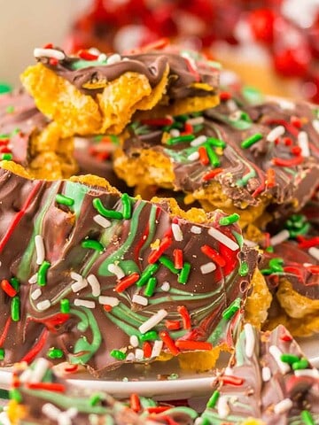 A plate of chocolate-covered treats topped with festive red, green, and white sprinkles. The background is decorated with a red, green, and white checkered cloth and scattered colorful candy spheres.