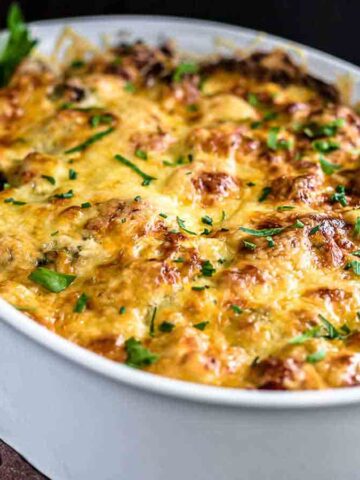 A baked casserole in a white oval dish, topped with golden melted cheese and garnished with fresh parsley. A fork and serving spoon are placed beside the dish on a brown cloth.