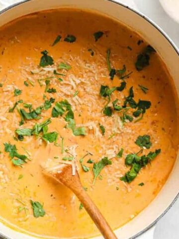 A pot of creamy tomato soup garnished with fresh parsley and grated cheese. A wooden spoon rests inside the pot. Next to the pot are small bowls containing extra cheese and parsley. The pot sits on a light-colored cloth on a marble surface.