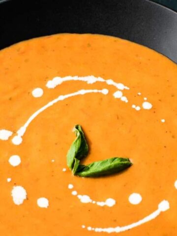 A black bowl filled with creamy orange soup garnished with a swirl of white cream and two fresh basil leaves, placed on a dark surface.