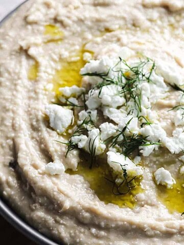 A bowl of hummus topped with crumbled feta cheese, a drizzle of olive oil, and fresh herbs. The hummus is served in a dark bowl placed on a wooden surface.