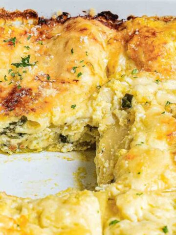 A close-up of a baking dish containing cheesy enchiladas. Some enchiladas are topped with melted cheese and herbs, while a section is cut out, revealing the creamy interior. The dish appears baked to a golden brown.