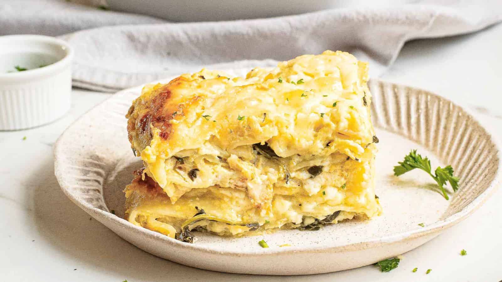A baked dish of zucchini lasagna with a golden-brown cheese topping. A wooden spatula lifts a square piece, revealing layers of zucchini, cheese, and spinach. The casserole dish is partly cut, showing more servings. Garnished with herbs.