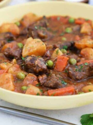 A bowl of hearty beef stew filled with chunks of beef, potatoes, carrots, and peas in a savory broth. A spoon is placed beside the bowl on the table, with some leaves and a carrot partially visible in the background.