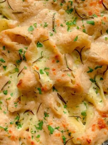 A close-up of a baked pasta dish in a round pan. The pasta is covered with a creamy sauce, garnished with chopped herbs, and is lightly browned on top. The pan is placed on a light-colored cloth.