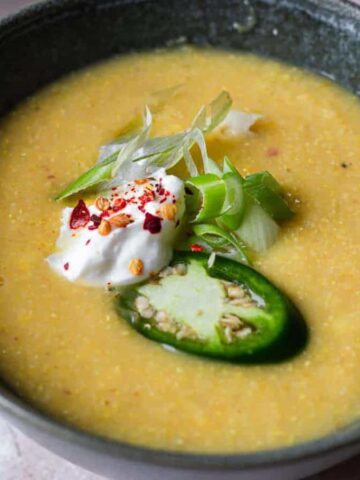 A bowl of creamy soup garnished with sliced jalapeño, a dollop of sour cream, chopped green onions, and red pepper flakes. A small plate of additional sliced jalapeños is visible in the background.