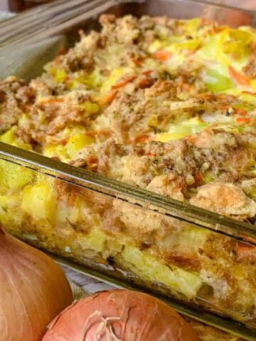 A glass baking dish holds a baked casserole topped with crumbly brown breading. The dish appears to contain chopped vegetables and possibly meat. Two whole onions and a green woven cloth are positioned beside it.