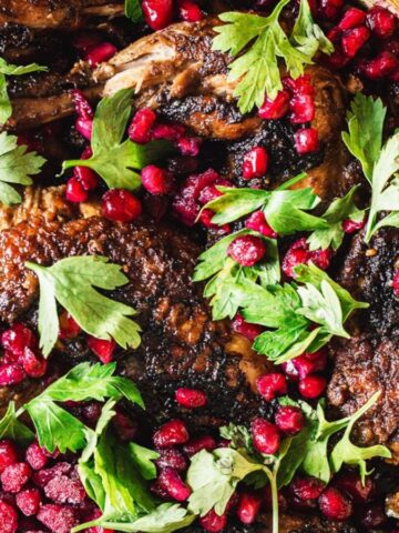 Cooked chicken in a pan garnished with pomegranate seeds and fresh parsley leaves. The dish has a rich, golden-brown color and is surrounded by a vibrant mix of ingredients.