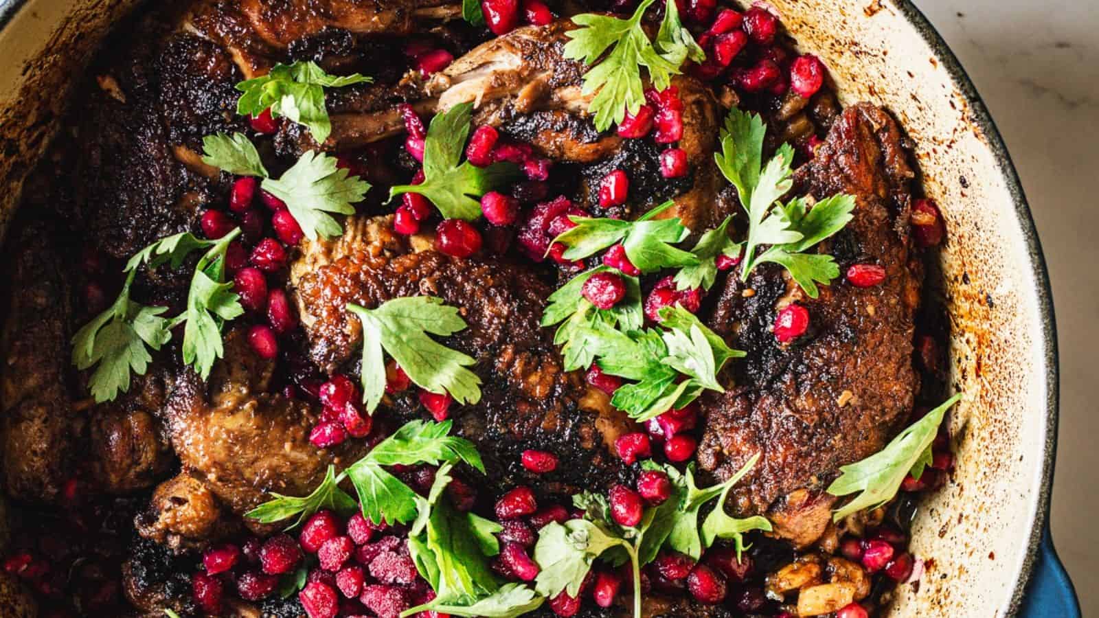 A close-up of a cooked dish featuring several pieces of meat topped with vibrant green parsley leaves and scattered pomegranate seeds. The dish is placed in a dark pot.