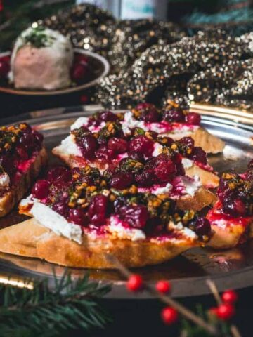 A platter of toasted bread topped with goat cheese, cranberries, and nuts sits on a festive table adorned with gold fabric and evergreen branches. A small bowl of dessert and a red drink are visible in the background.