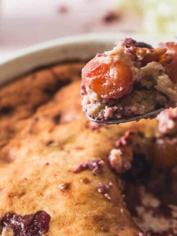 A close-up of a baked dessert in a round dish with a portion scooped out. The dessert appears golden brown with visible cherries and a moist texture. A spoon holds a serving of the dessert above the dish.