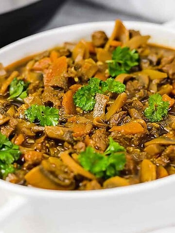A white ceramic dish filled with beef stew, featuring chunks of beef, sliced mushrooms, and diced carrots, garnished with fresh parsley. A spoon is placed beside the dish on a white surface.