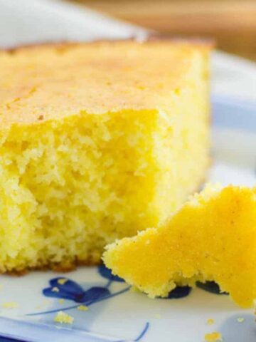 A slice of cornbread on a white and blue plate with two small crumbles beside it. The cornbread is golden yellow with a slightly crispy edge, sitting on a light background.
