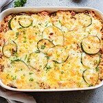 A rectangular baking dish holds a ground beef zucchini and rice casserole topped with melted cheese and slices of zucchini. The dish is garnished with chopped parsley, resting on a light-colored surface.