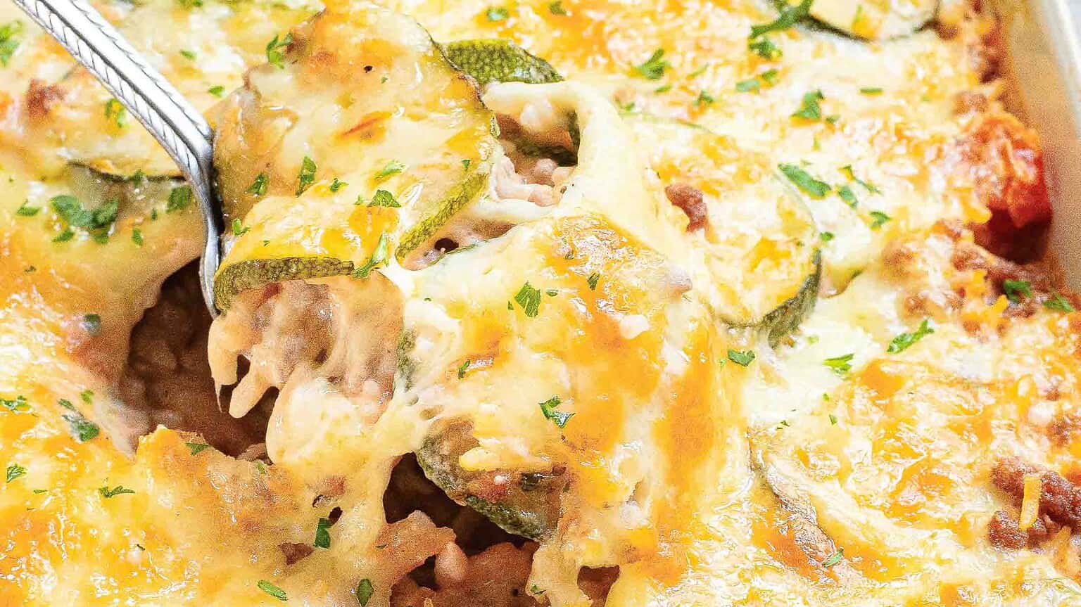 A close-up of cheesy baked zucchini with meat, parsley, and golden-brown melted cheese, served in a dish with a fork.