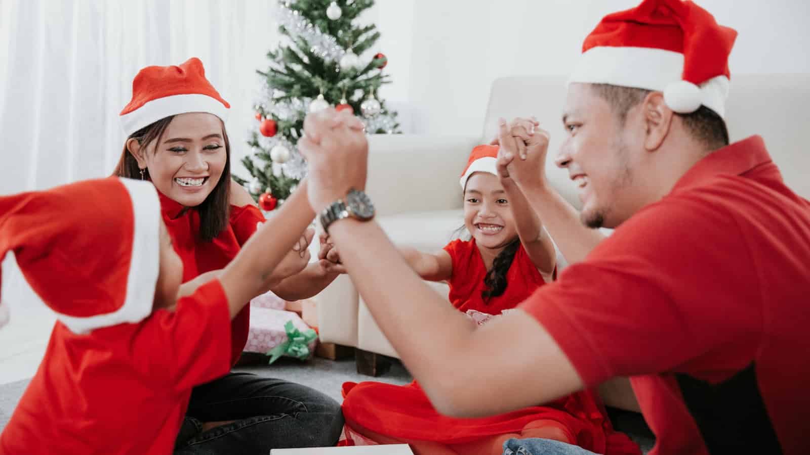 A family of four, embracing Christmas Eve traditions, sits on the floor wearing red Santa hats and clothes. They hold hands in a circle, smiling joyfully as kids often do. In the background, a decorated Christmas tree stands near a cozy sofa.