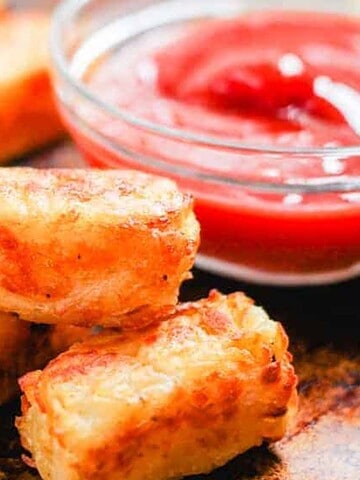 A batch of golden-brown tater tots is arranged on a tray. A small glass bowl filled with red ketchup stands next to them. The tots are crispy and appear freshly baked.