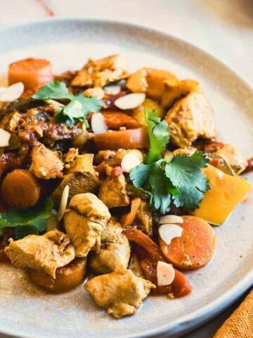 A plate of stir-fried chicken with carrots, bell peppers, and almonds garnished with cilantro. A gold fork is on the plate, and an orange napkin is beside it on a marble surface.