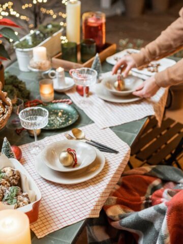 A festive table is set with candles, cookies, and decorated plates. A person showcases tricks for special holiday breakfasts while placing a bowl on the table. Nearby, a large fluffy dog sits wrapped in a plaid blanket. Poinsettias and greenery enhance the cozy holiday atmosphere.