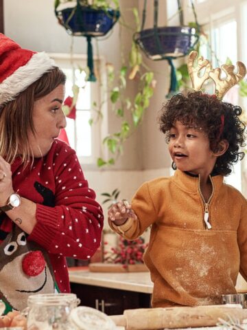 An adult and a child are in the kitchen, wearing festive clothing. The adult dons a Santa hat, holding a ball of dough ready for perfect pies, while the child wears a crown and excitedly points at something. Baking ingredients are scattered across the countertop, setting the scene for holiday delights.