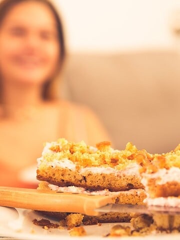 Close-up of two slices of crumb cake, an unexpected American comfort food, with creamy layers on a wooden board. In the blurred background, a person is sitting on a couch.