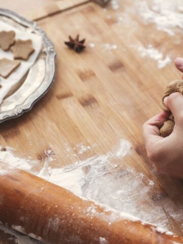A person kneads dough on a wooden surface next to a rolling pin. Cookie cutters and flour are scattered around, evoking classic holiday recipes. A tray with cut-out shapes, including trees and circles, adds a modern twist. An anise seed is on the table.