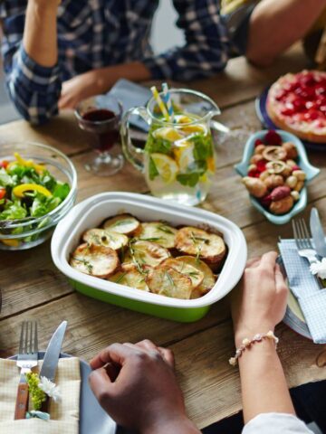 A group of people seated around a wooden table with a variety of dishes, including an intriguing array of strange pot luck dishes like unique casseroles and exotic salads. Sliced bread, pie, drinks, and a wine bottle complete the scene, suggesting a casual gathering or meal.