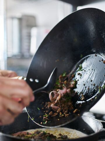 A person cooking in a kitchen is skillfully using a spatula to slide sautéed vegetables from a wok into a frying pan, effortlessly repurposing holiday leftovers. The background reveals an array of kitchen equipment and utensils.