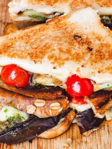 A grilled sandwich is cut in half on a wooden board. It contains eggplant, cherry tomatoes, zucchini, melted cheese, and pesto. A bowl of green pesto sauce is next to the sandwich, and there is a knife on the side.