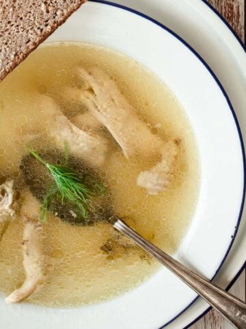 A bowl of clear chicken soup with shredded chicken pieces and a sprig of dill. A slice of rye bread rests on the edge of the bowl. A spoon is placed in the soup. The setup is on a rustic white wooden surface.
