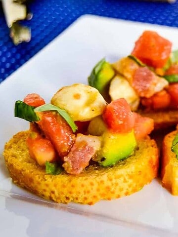 Three bruschetta toasts topped with diced tomatoes, mozzarella balls, avocado, basil, and pieces of cured meat are arranged on a white square plate placed on a blue textured surface.