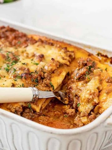 A baked lasagna in a rectangular white dish with a serving spoon placed inside. The top is golden brown with melted cheese and garnished with herbs. A portion of the lasagna has been served, revealing layers of pasta, meat, and cheese.