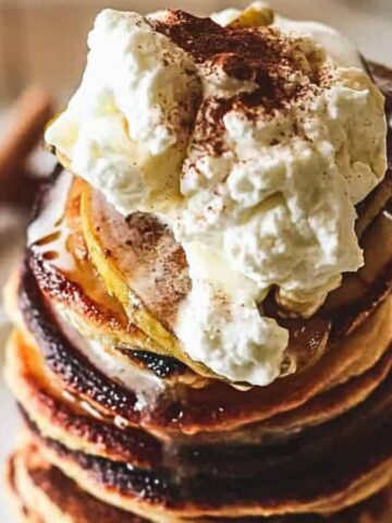 A stack of pancakes sits on a white plate, topped with a generous dollop of whipped cream and sprinkled with cinnamon. A drizzle of syrup runs down the sides. In the background, there's a blurred view of a cinnamon stick and a bowl.