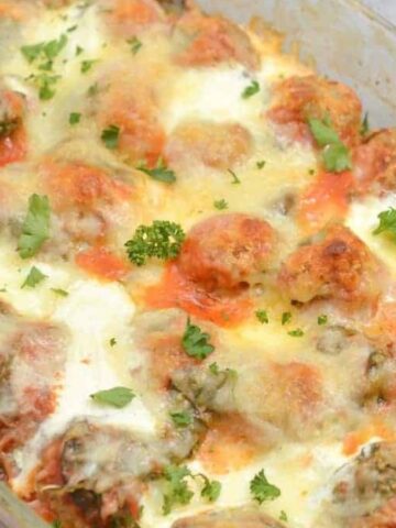 A baked dish of cheesy meatballs topped with melted cheese and garnished with parsley, served in a glass baking dish. The dish is placed on a gray textured surface.