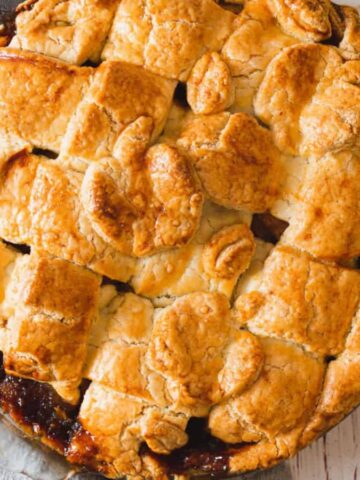 A freshly baked apple pie with a lattice crust is on a wooden surface. A gray cloth is partially visible on the left, and a sliced green apple is on the right, next to a filled bowl. The pie has a golden-brown, flaky appearance.