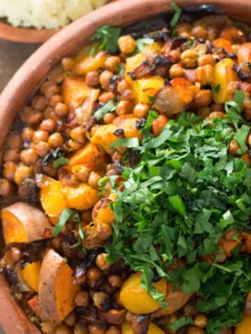 A bowl of vegetable stew with chickpeas, carrots, and bell peppers, garnished with chopped herbs. A small bowl of chopped herbs and a side dish with grains are on a wooden surface nearby.