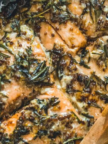 Close-up of cooked salmon fillets in a pan with a savory herb and seasoning mixture. A wooden spoon rests at the edge, coated with the pan's contents. The dish features visible rosemary, mustard seeds, and a glossy sauce.